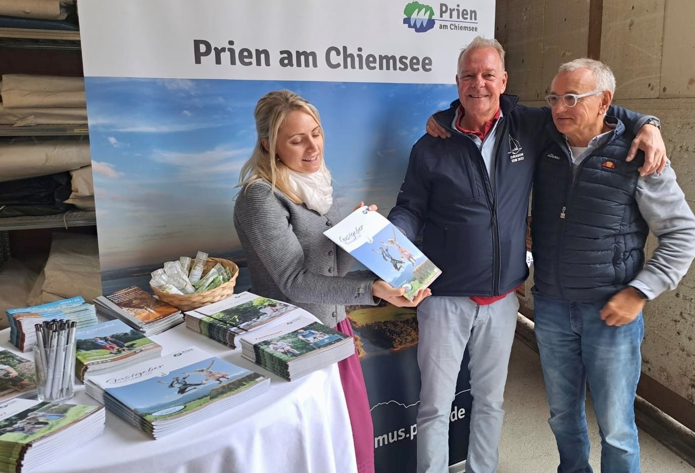 Infostand auf dem Gelände des Chiemsee Yacht Club e.V.: (v.l.n.r.) Die Mitarbeiterin des Tourismusbüros Prien Stefanie Sattlberger präsentiert den IDM-Besuchern aus Berlin und Düsseldorf das Gastgeberverzeichnis der Marktgemeinde Prien a. Chiemsee. Die 56 Seiten lange Broschüre zeigt eine breite Angebotspalette auf. Neben dem umfangreichen Gastgeberverzeichnis ist in dem ansprechenden Heft eine Vielzahl an Veranstaltungen, Führungen und Aktionen aufgeführt.Bildrechte: Markt Prien a. Chiemsee